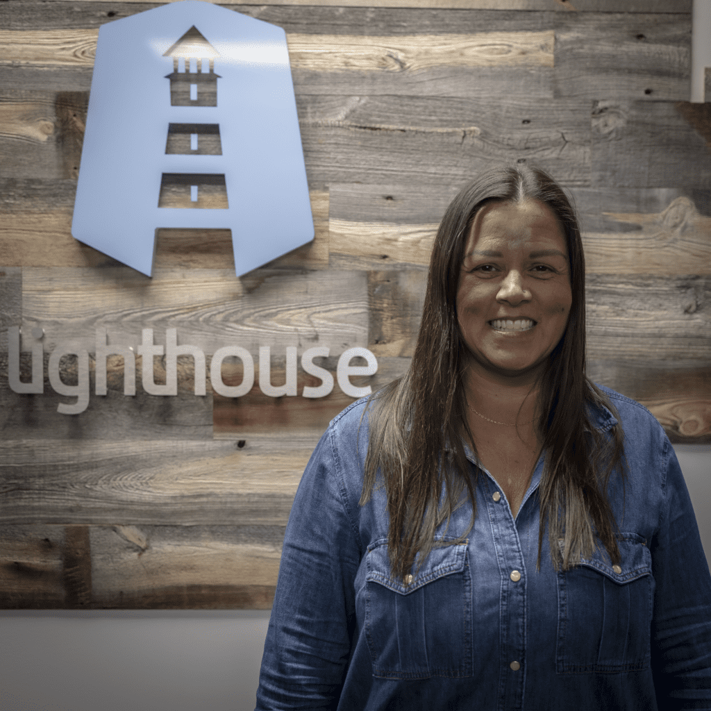 Office Manager, Tammy Stang smiling in front of the Lighthouse sign on wooden wall