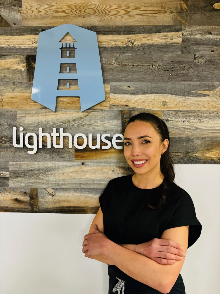 RMT, Stacy Salvati standing with crossed arms in front of the lighthouse logo on wooden wall